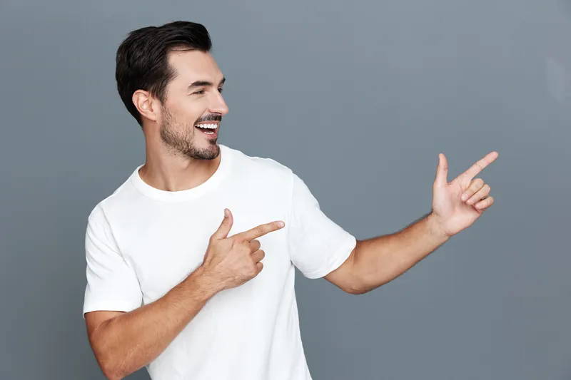 Smiling man points to the side while looking at the camera, dressed in a white t-shirt with a joyful expression, standing against a solid gray background.