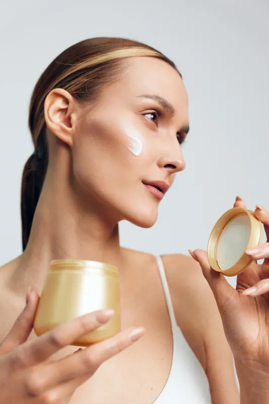 Young woman with clear skin applying face cream from a jar. Close-up portrait on a light background. Concept of skincare, beauty, self-care, and moisturizing routine.