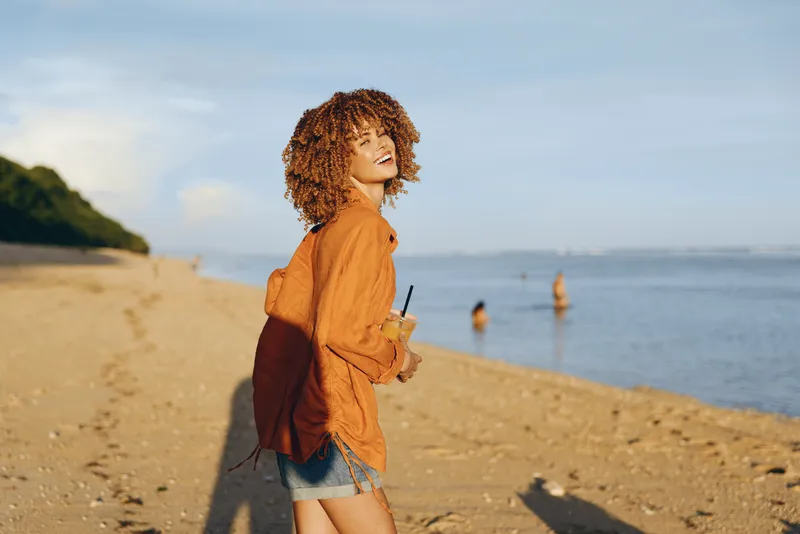 Woman with curly hair on beach near sea, sunlight on face, outdoors casual denim jacket shorts, laughing in profile, sandy shore and distant waves, calm summer evening