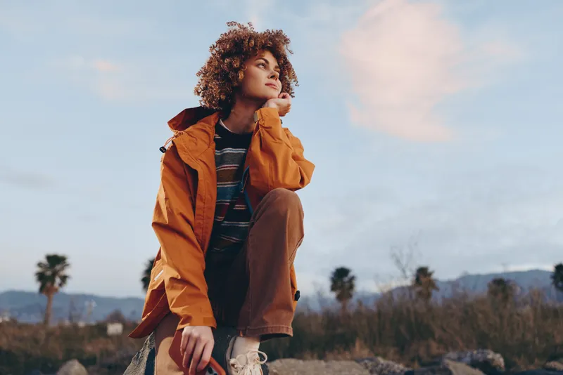 Woman lifestyle outdoors in autumn light, curly hair in an orange jacket, standing on a rocky field, open space, calm thoughtful pose, soft wind, cloudy sky, distant palm trees