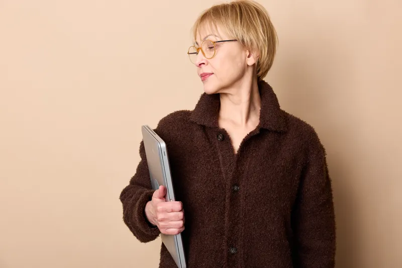 Woman tablet glasses portrait of a blonde adult holding a tablet, looking sideways with calm focused expression, indoor beige background, casual fur coat, technology communication