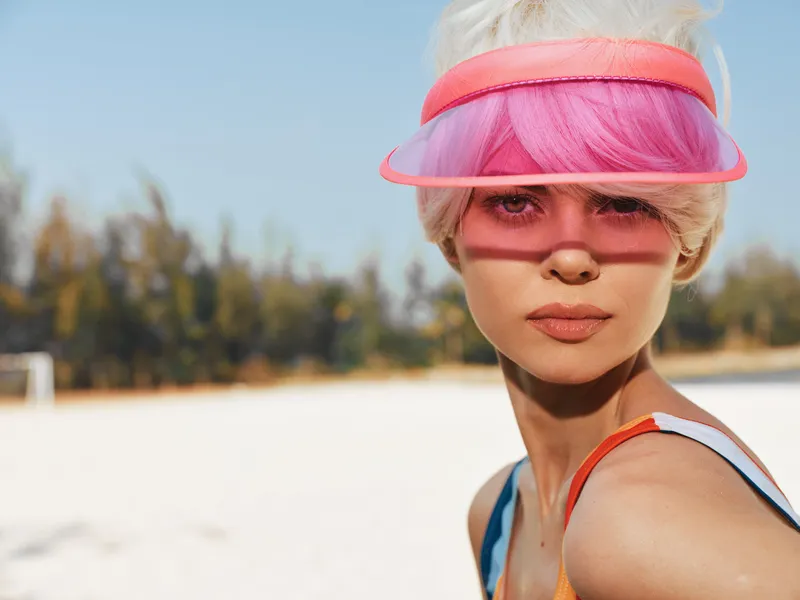 Portrait of a stylish young woman on a bright beach wearing a pink visor, vibrant hair, and a summer outfit. Warm light highlights her calm, confident gaze.