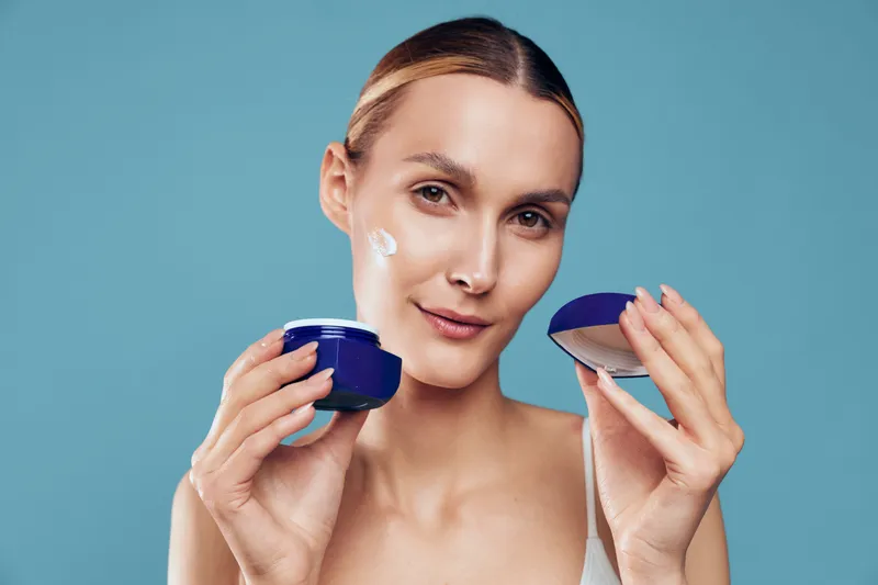 Young woman with clear skin applying white face cream from blue jar on her cheek against blue background. Concept of skincare, beauty routine, wellness, and self-care.