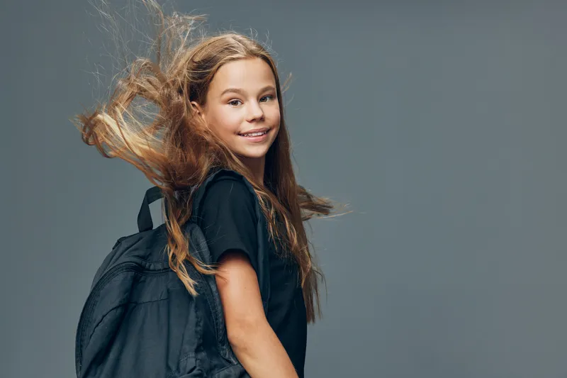 Girl with long hair smiling and carrying backpack against grey background. Child with casual clothing and hair blowing in wind for school or travel concept.
