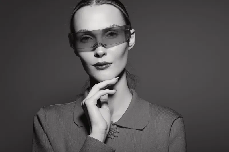 Elegant white woman with pale skin wears oversized transparent glasses and textured jacket, posing thoughtfully against a dark background.