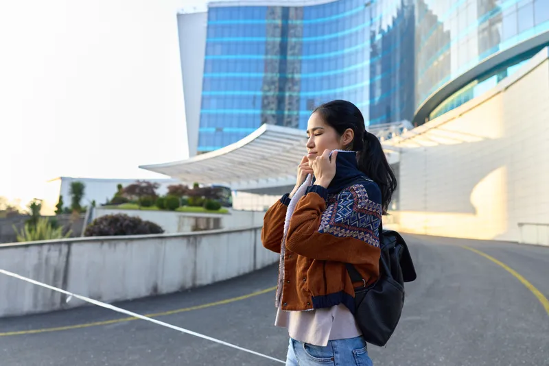 Woman on a phone in an urban cityscape, outdoor scene with modern architecture, brown jacket and backpack, casual dress, street curve, daytime commute, candid lifestyle moment