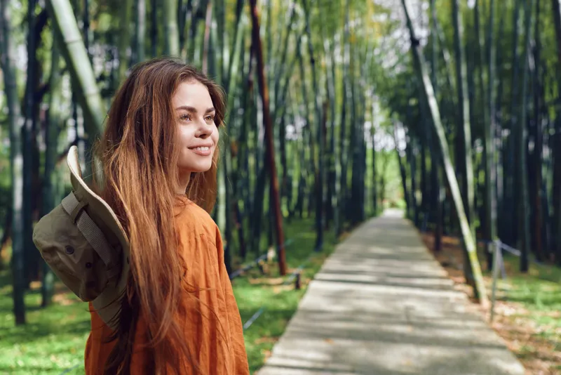 Woman with backpack and smile on a paved path through a bamboo forest, nature travel portrait of a young girl walking outdoors, enjoying lush greenery and a peaceful scenic journey.