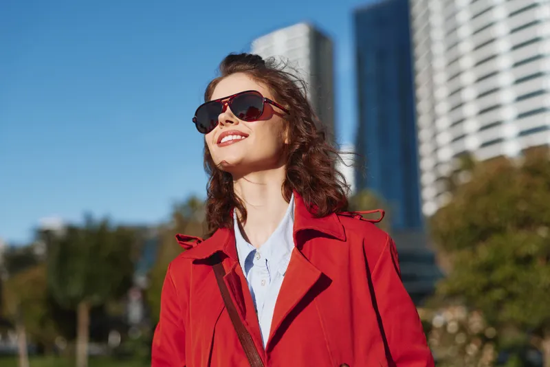 A stylish woman in a red coat and sunglasses walks outdoors in an urban setting, smiling under bright afternoon light, capturing confident street fashion and sunny vibes.