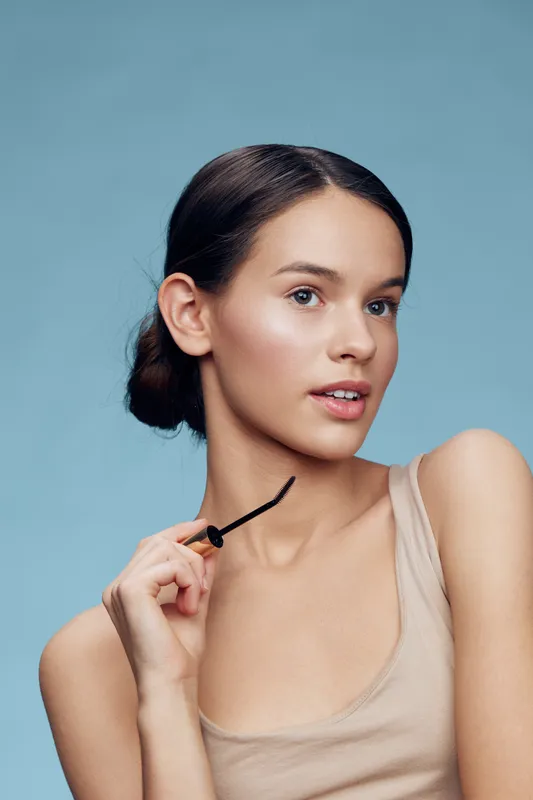 Young woman with a neutral expression holding a hair accessory, dressed in a nude top, against a soft blue background, showcasing a minimalist and modern aesthetic