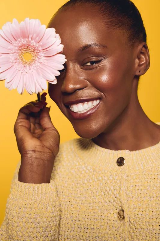 Smiling black woman with dark skin holds pink flower near face against yellow background. Concept of natural beauty, happiness, and skincare with joyful female portrait in studio lighting.