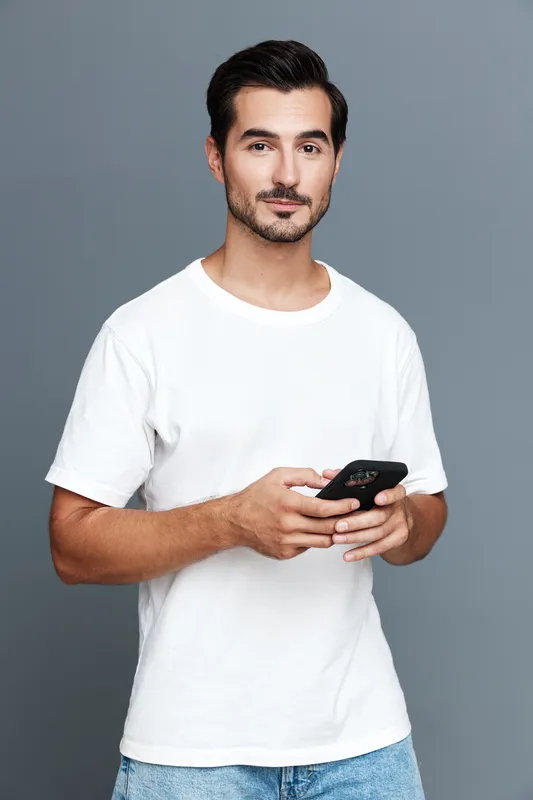 Casual smiling man in a white t-shirt holding a smartphone, looking confidently at the camera, set against a neutral gray background. Modern lifestyle concept.