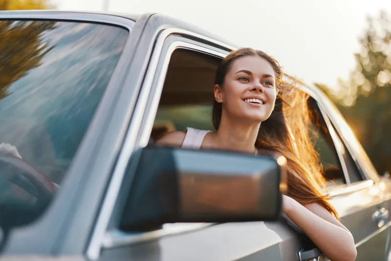 Young woman enjoying a road trip, smiling joyfully with her head out of the car window, surrounded by greenery during sunset, capturing the essence of freedom and adventure.