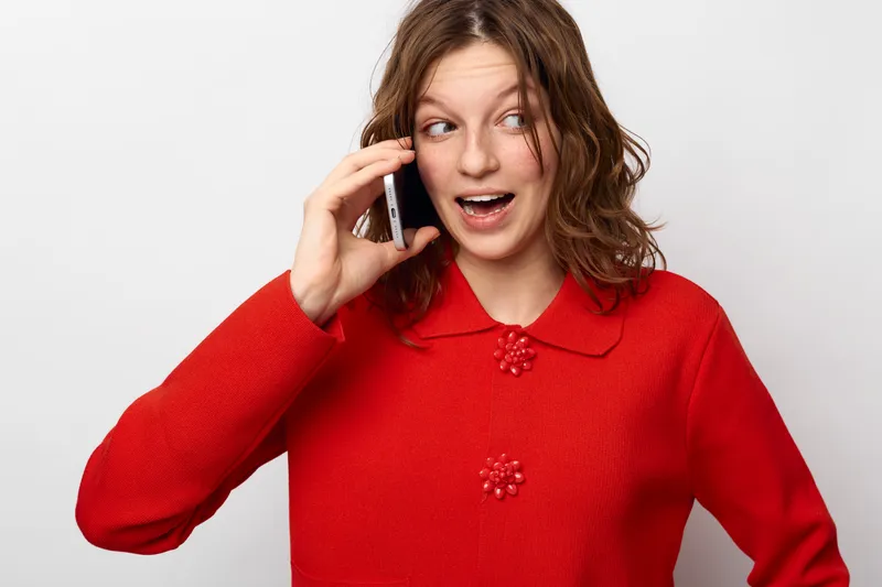 Young woman in red sweater talks on smartphone with surprised expression. Isolated on white background. Concept of communication, technology, emotions, and casual lifestyle.