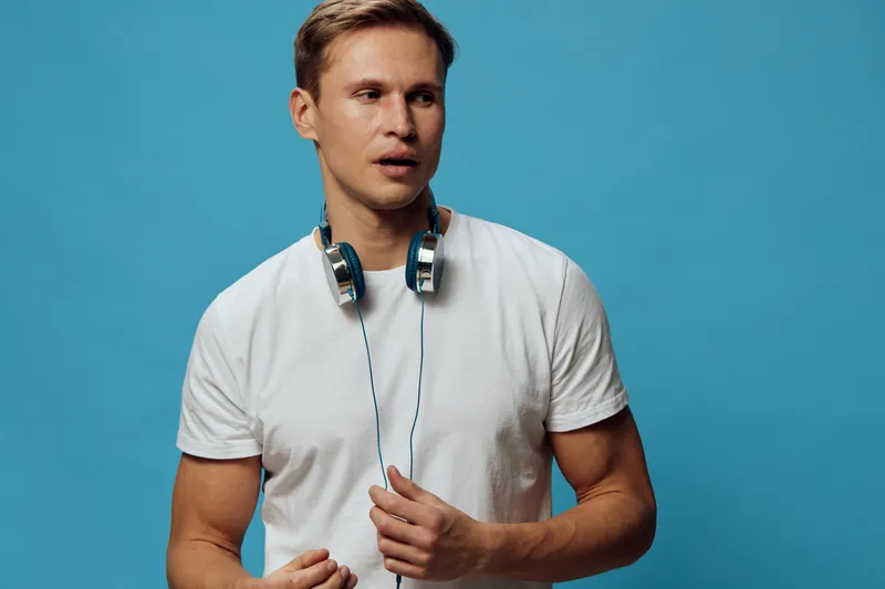 Young man with headphones around neck wearing casual white t-shirt, looking to the side with surprised expression, standing against solid blue background, studio shot, casual style, men lifestyle conc