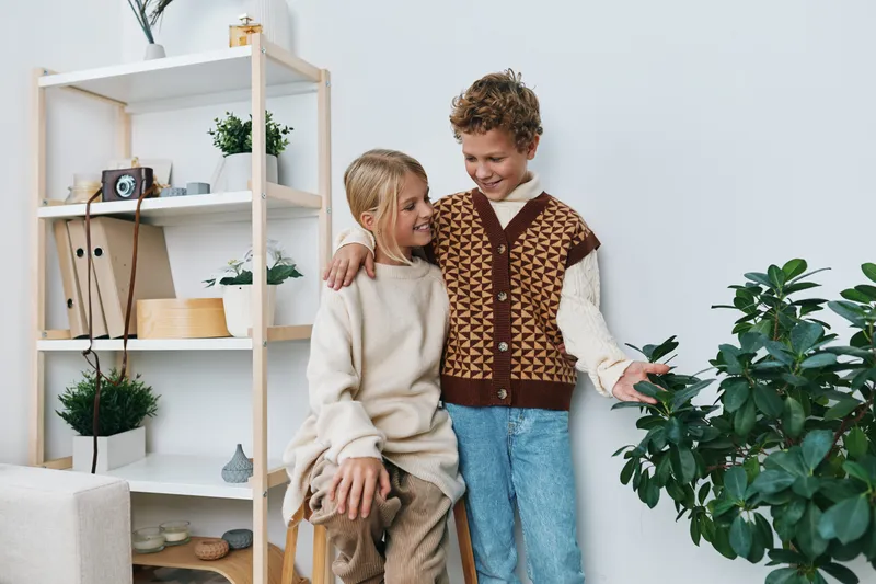 Two cheerful children share a warm moment indoors, wearing cozy knitted sweaters, standing near a wooden shelf and lush houseplants in a bright, modern room.