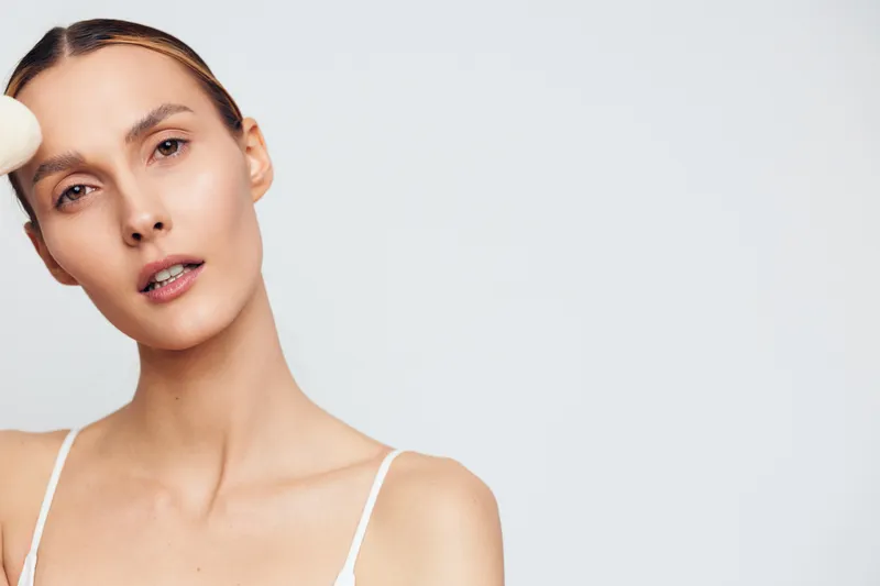 Young woman with clear skin applying white face cream with a cotton pad on her forehead. Minimalist background emphasizes skincare and beauty routine. Soft natural lighting highlights texture.