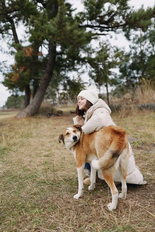 Woman in a warm coat kneels beside a large dog in an open park, embracing the loyal companion on a chilly autumn day, amid serene nature