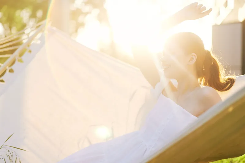 Cheerful young woman relaxing in a hammock, illuminated by golden sunlight, wearing a white dress, in a serene outdoor setting with a soft green background
