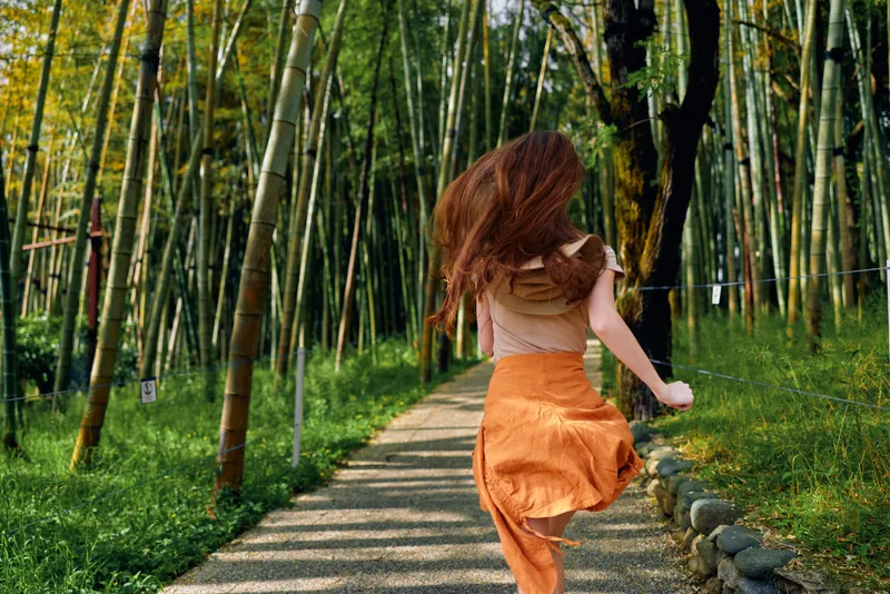 Woman running along a bamboo forest path in nature, wearing an orange skirt, long red hair flowing, back view motion on sunlit trail with green foliage and peaceful atmosphere