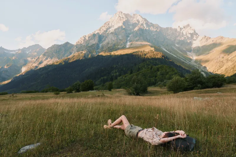 A woman in casual clothes lies on grass in a mountain meadow, relaxing under a partly cloudy sky with tall peaks in the background, enjoying nature and lifestyle outdoors.