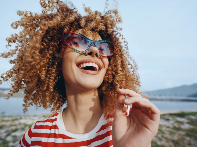 Joyful young woman with curly hair and colorful sunglasses laughing on a sunny beach, striped shirt, wind in her hair, bright sky, carefree moment of summer happiness.