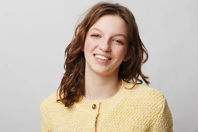 Smiling young woman with brown hair wearing a yellow knitted sweater looks at camera on grey background. Portrait of happy female model with natural makeup and casual style in studio lighting.