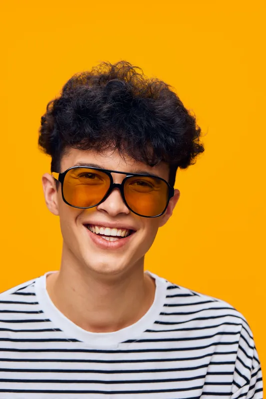 smiling man with curly hair wearing yellow sunglasses and striped shirt against bright orange background, expressing happiness and youthfulness