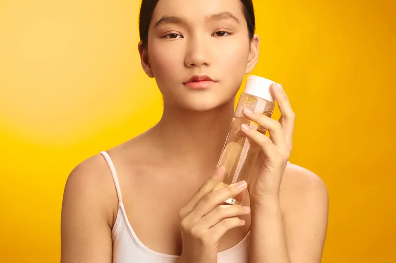 asian woman holding a bottle against a warm yellow backdrop, showcasing skincare product with calm expression and clean aesthetic, ideal for beauty, wellness and cosmetics campaigns.