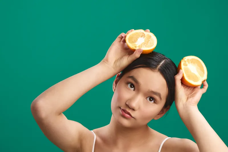 Asian woman holds citrus halves against a vibrant green backdrop, tilting her head with a soft smile to convey a fresh, energetic mood suitable for lifestyle and wellness visuals.