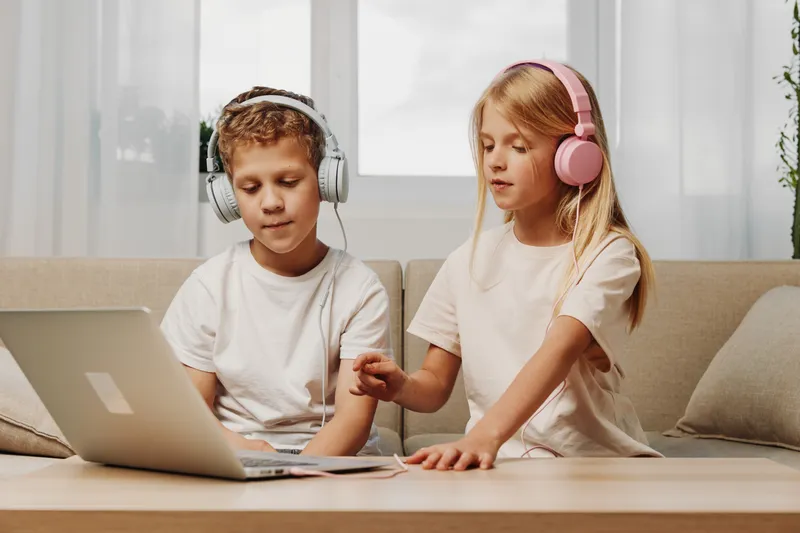 Two children with headphones use laptop on wooden table in cozy living room with soft natural light for study and entertainment.