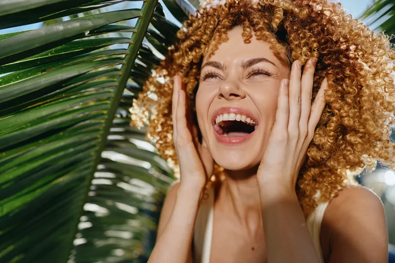 joyful woman with curly hair laughing outdoors near tropical leaves under bright sunlight, experiencing happiness and natural beauty in summer.