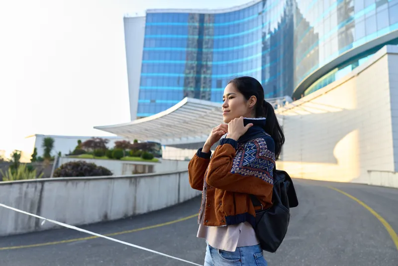 Woman in a casual brown jacket with a backpack stands on an urban walkway beside modern glass buildings, adjusting her collar, conveying street style, travel readiness, and a forward looking mood