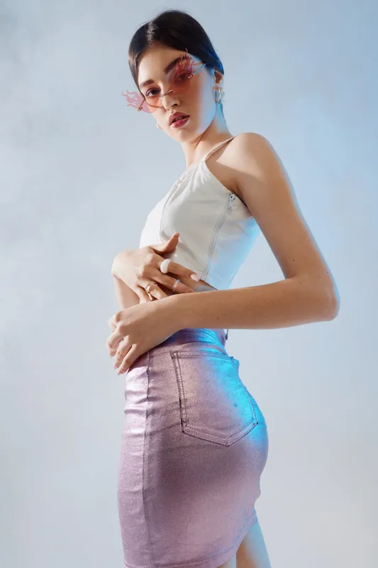 Young woman in a white top and shiny pink skirt poses confidently wearing fashionable sunglasses. Studio shot with soft lighting on a light background. Concept of beauty fashion style.