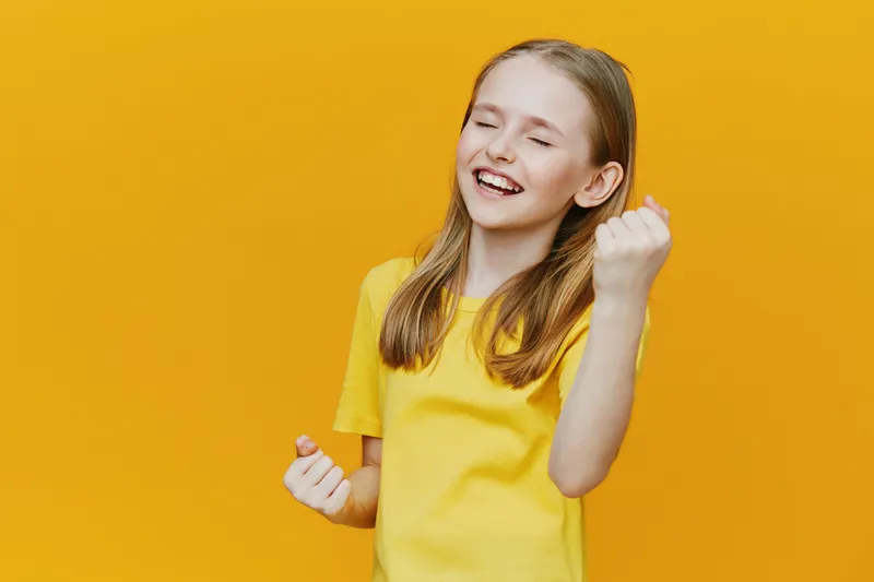 Happy young girl celebrating success with a joyful expression against a vibrant yellow background, wearing a bright yellow shirt