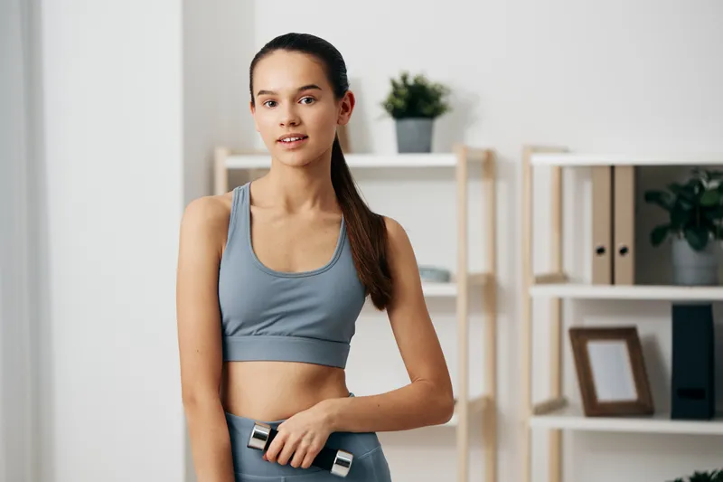 Woman in athletic wear holding a dumbbell during a home workout, bright room with shelves, plants and framed art, showcasing personal training, fitness routine, and concentration.