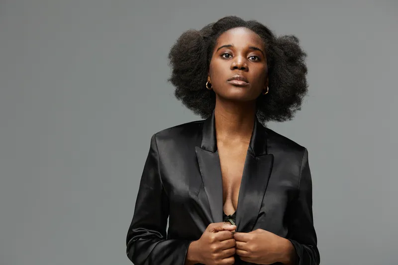 Elegant young woman with natural hair in a black blazer, exuding confidence against a neutral gray background, ideal for fashion and empowerment themes