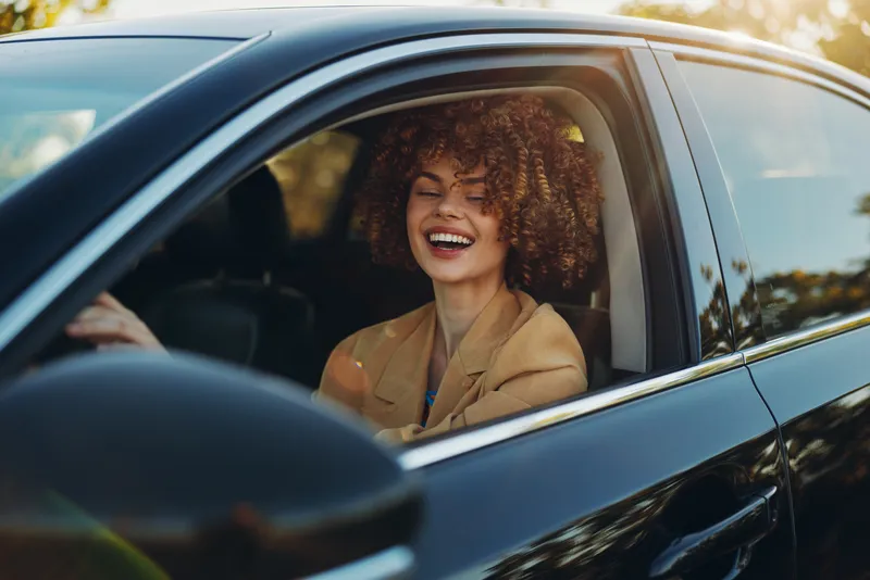 A young woman with curly hair laughs in the driver's seat of a sunlit car, enjoying a carefree moment as warm light filters through the window and frames her joyful expression, and she glances toward