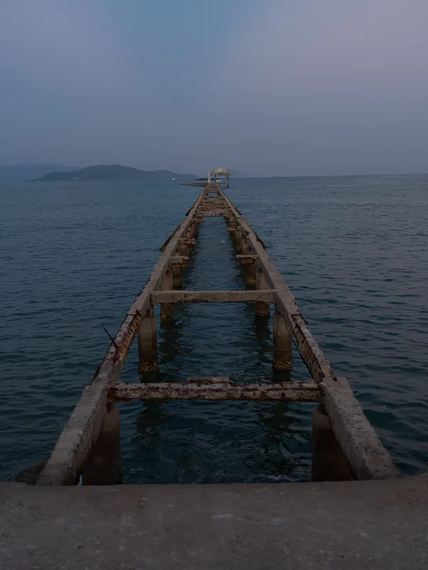 Abandoned rusty pier extends into calm ocean under dusk sky, creating a serene and moody atmosphere with distant islands on the horizon.