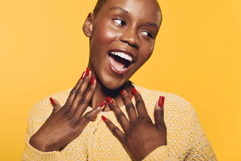 Happy dark skinned African woman with short hair and red nails smiles widely, wearing yellow sweater on yellow background. Concept of joy, beauty, and positive emotions in portrait.