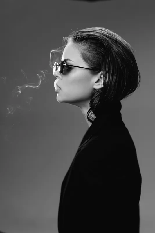 Black and white profile of a stylish woman in sunglasses exhaling smoke, wearing a dark jacket.