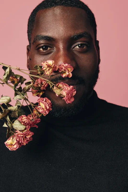 African man with dark skin holding dried pink flowers close to face in creative conceptual visual style. Portrait with black turtleneck and soft pink background for artistic expression.