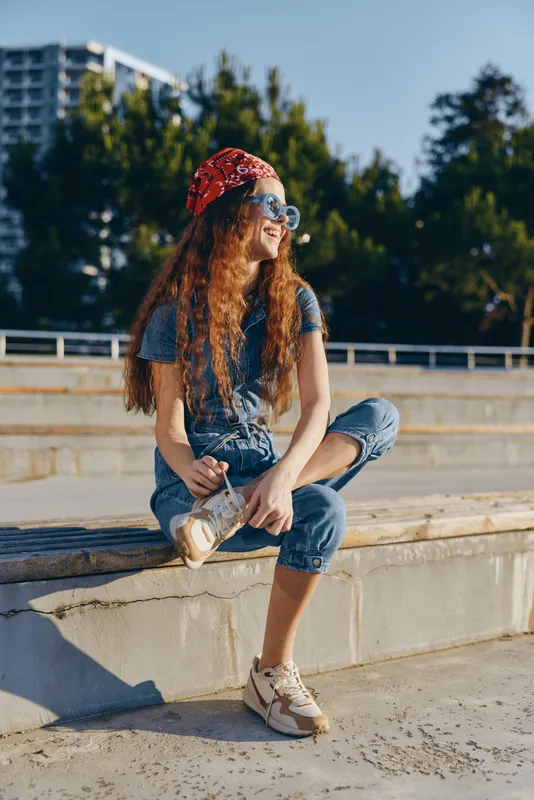 Lifestyle woman in modern boho-western denim street style sitting outdoors in warm sunlight wearing sunglasses and a red bandana, film color effect enhancing the casual vibe
