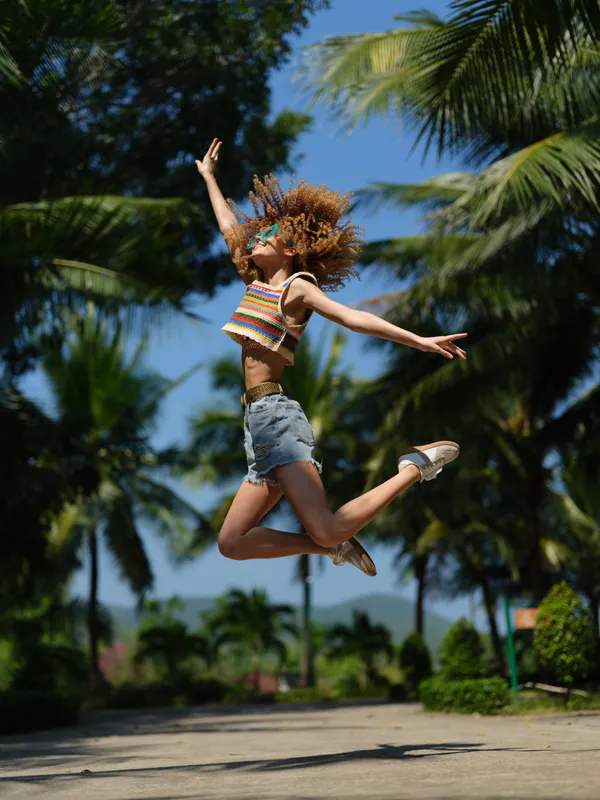 joyful woman jumping outdoors with colorful top and denim shorts, palm trees background, summer energy and freedom in bright daylight