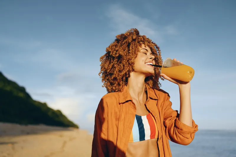 Smiling woman outdoors on a sunny coast, curly hair, drinking from a bottle, wearing a brown jacket and striped shirt, sea view and cliffs behind, natural light, relaxed posture, calm smile