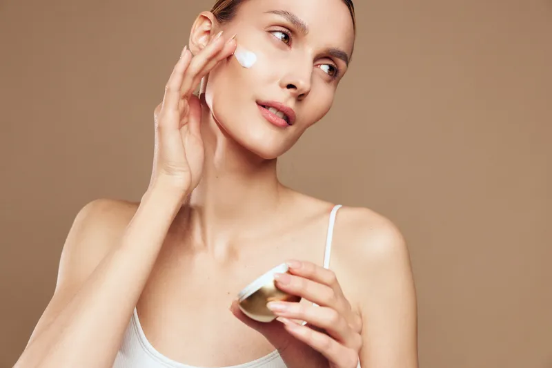 Young woman applying face cream on cheek, natural skincare routine, smooth skin, soft lighting, beige background, calm expression, beauty care concept