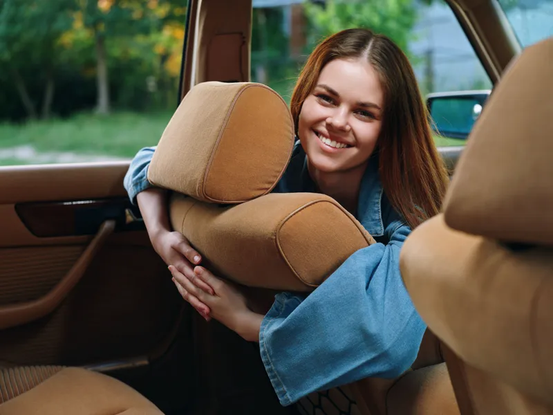 Young woman smiling in a vintage car, wearing a denim jacket and leaning on a tan pillow, exuding happiness and warmth in a natural outdoor setting.