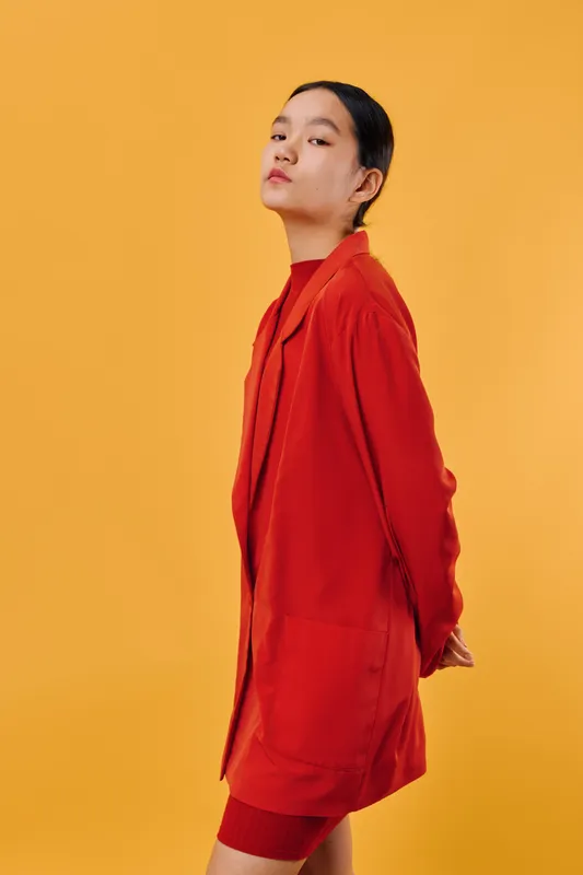 Young woman in red jacket posing confidently with hands behind back on bright yellow background. Minimalist fashion portrait with bold colors and modern style for editorial or advertising.