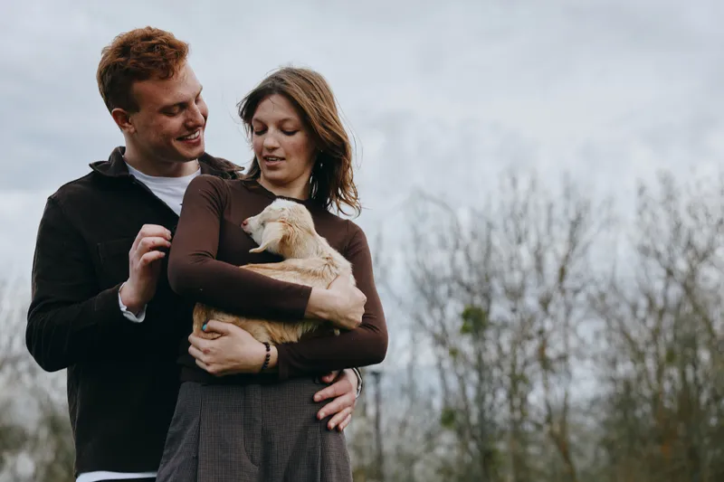 Couple holds a goat kid in their arms outdoors, sharing affection and a joyful moment, showcasing rural lifestyle and tender bond in a natural, warm scene.