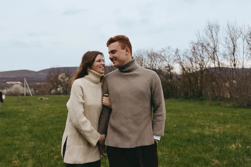 A couple in cozy sweaters share a warm moment in a grassy field, smiling as they look at each other, showcasing affection and everyday intimacy in a relaxed lifestyle scene.