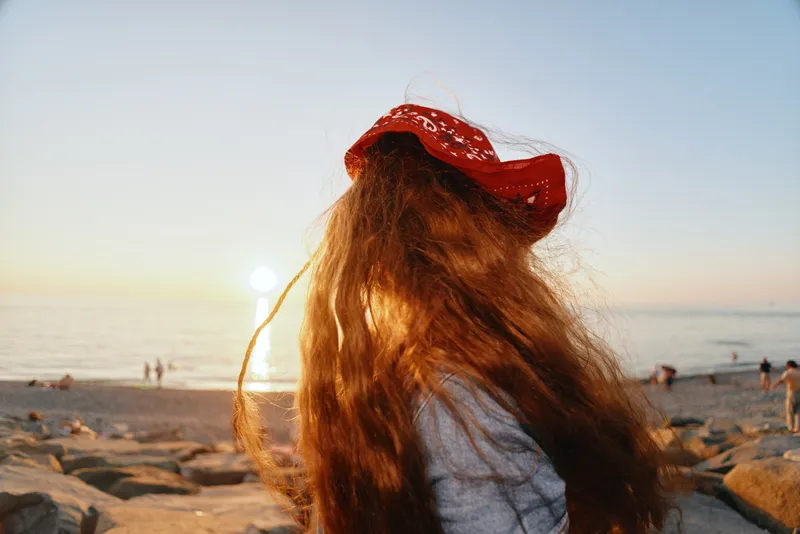 Woman with long hair and red bandana enjoying sunset on rocky beach, calm sea, warm light, serene atmosphere, summer evening relaxation.